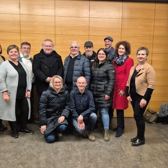Eine Gruppe von Menschen, sowohl Männer als auch Frauen, posiert für ein Foto vor einer Holzwand. Sie alle lächeln und tragen Winterkleidung.