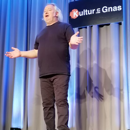 Ein Mann steht auf der Bühne, spricht und hat die Arme weit ausgestreckt. Hinter ihm befindet sich eine Kulisse mit einem Schild, das 'Kultur = Gnäs' besagt.