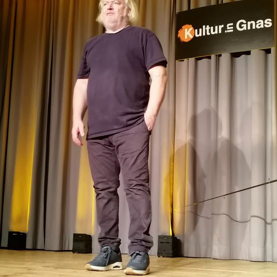 Ein Mann steht auf der Bühne, trägt ein lilafarbenes Hemd und eine Hose, hinter ihm ein Schild mit der Aufschrift 'Kultur. Gnas'.