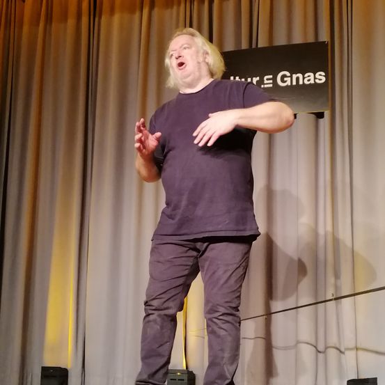 Ein Mann steht auf der Bühne und singt mit offenem Mund. Er trägt ein lilafarbenes T-Shirt und eine Hose. Hinter ihm ist ein Vorhang hochgezogen. Ein schwarzes Schild mit weißem Text befindet sich über ihm.