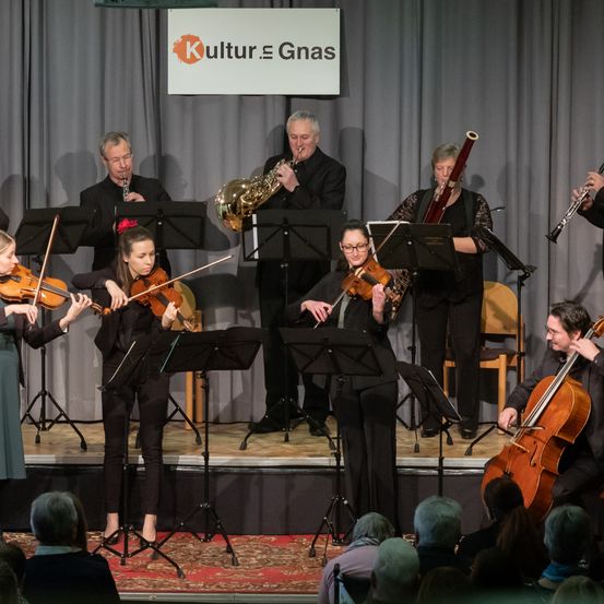 Eine Gruppe von Musikern spielt auf der Bühne mit Violinen, Celli und Blasinstrumenten. Dahinter befindet sich ein Banner mit 'Kultur E Gnas'. Das Publikum sitzt vor der Bühne.