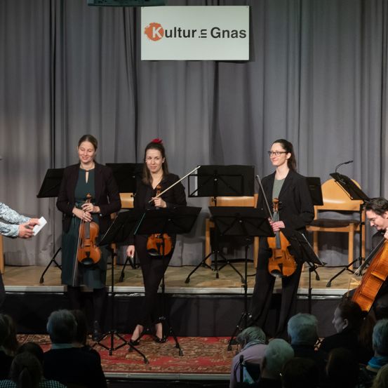 Vier Musiker stehen mit Violinen und Cello auf der Bühne und spielen vor einem Publikum. Hinter ihnen befindet sich ein Banner mit der Aufschrift 'Kultur. Gnas.' Die Bühne ist mit grauen Vorhängen und einem Teppich dekoriert.