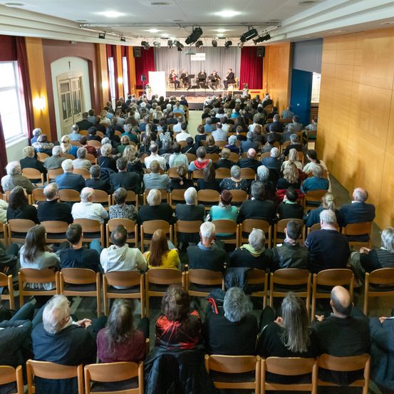 Ein großes Publikum sitzt in Stuhlreihen und beobachtet eine Aufführung auf der Bühne. Einige Musiker spielen Instrumente. Der Raum hat Holzwände und eine hohe Decke mit Beleuchtung.
