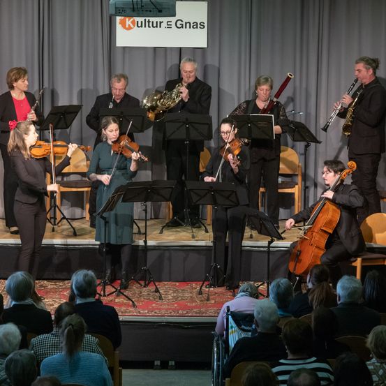 Ein Musikensemble tritt auf der Bühne auf, mit Geigern, Cellisten und Blechbläsern. Dahinter zeigt eine Leinwand das Logo von Kultur Gnäs. Das Publikum sitzt aufmerksam und ist in die Aufführung vertieft.