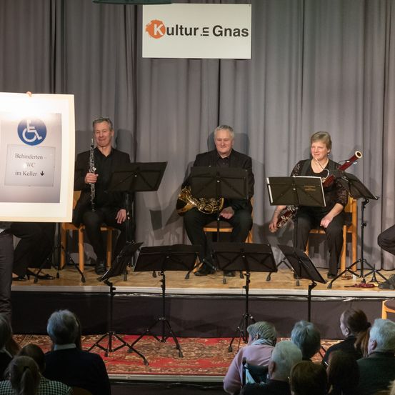 Ein Musikauftritt auf der Bühne mit vier Personen, darunter ein Klarinettist, ein Hornist und ein Fagottist, vor einem Publikum. Die Bühne ist mit einem Banner geschmückt, das 'Kultur & Gnas' besagt.