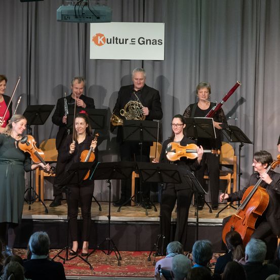 Eine Gruppe Musiker tritt mit verschiedenen Instrumenten, darunter Violinen, Celli und Blechbläser, auf der Bühne vor einem Publikum auf. Ein Schild mit dem Wort 'Kultur' ist sichtbar.