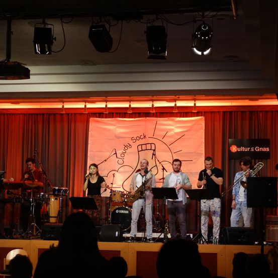 Eine Musikband tritt auf der Bühne vor roten Vorhängen auf. Sie wird von Bühnenlichtern beleuchtet. Vor ihnen befindet sich ein Banner mit der Aufschrift 'Cloudy Sock'. Das Publikum sieht zu.