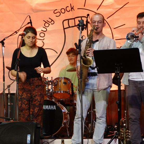 Eine Frau singt in ein Mikrofon auf der Bühne, begleitet von einem Mann, der Saxophon spielt, und einem anderen, der Trompete spielt. Hinter ihnen spielt ein Schlagzeuger ein Schlagzeug. Die Bühne hat einen Hintergrund mit dem Text 'Cloudy Sock'.