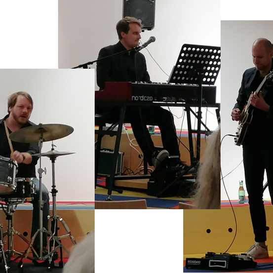 Drei Männer spielen auf der Bühne, einer spielt Gitarre, ein anderer Schlagzeug und der dritte am Keyboard.