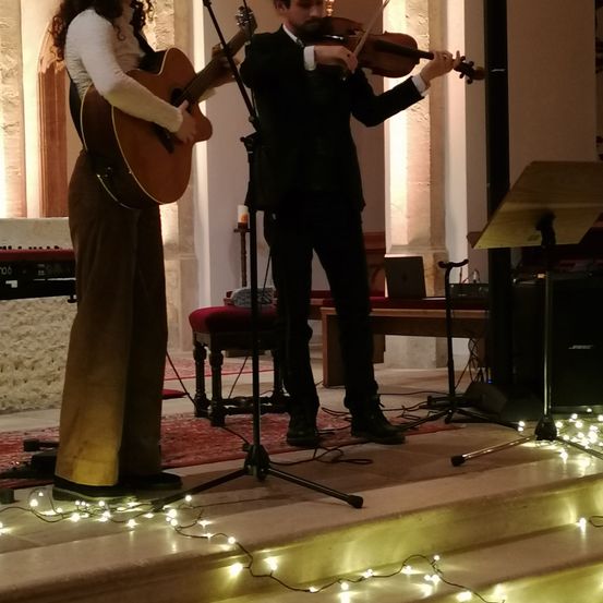 Zwei Musiker treten auf der Bühne auf. Eine Frau spielt Gitarre und ein Mann Geige. Sie sind von Lichterketten beleuchtet.