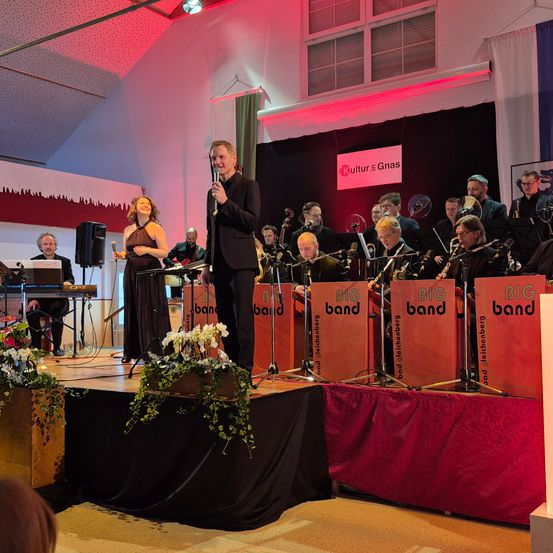 Eine Band auf der Bühne, mit einer Frau und einem Mann davor. Dahinter spielt eine große Gruppe von Musikern Instrumente. Es gibt Blumen und einen Lautsprecher auf der Bühne. Ein Banner hängt an der Wand.