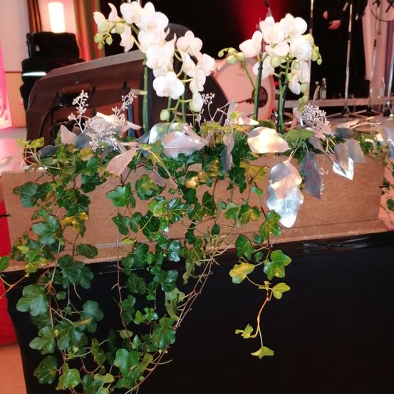 Eine Nahaufnahme einer dekorativen Anordnung mit weißen Orchideen, grünen Blättern und silbernen Bändern auf einem Tisch. Die Szene spielt in einem Raum mit einer Bühne und einem Schlagzeug im Hintergrund.