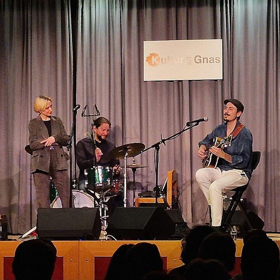 Drei Musiker treten auf der Bühne auf. Eine Frau steht links, ein Mann spielt Gitarre rechts und ein anderer Mann Schlagzeug in der Mitte. Ein Schild mit der Aufschrift Kultur Gnas ist hinter ihnen sichtbar.