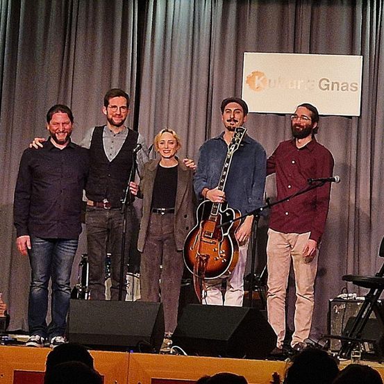 Fünf Musiker stehen lächelnd auf der Bühne, mit einem Banner im Hintergrund. Sie haben eine Gitarre, Mikrofone und Lautsprecher. Der Mann rechts trägt eine Brille.