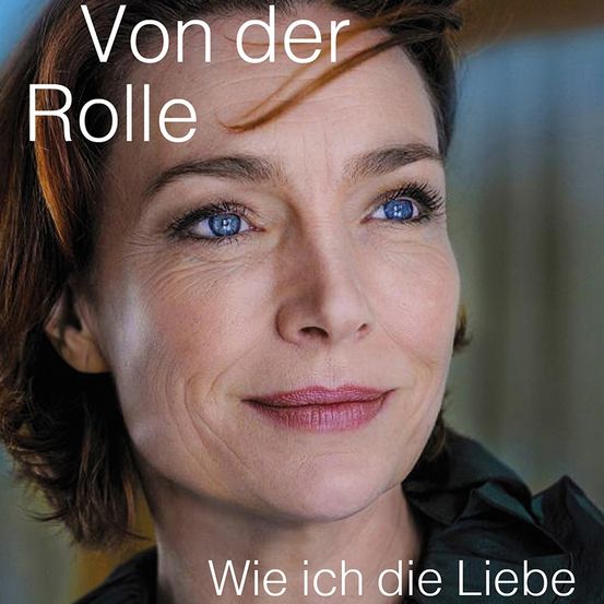 Eine Frau mit kurzen braunen Haaren und blauen Augen lächelt in die Kamera. Sie trägt ein schwarzes Oberteil. Über ihrem Kopf steht weißer Text, der 'Von der Rolle' lautet. Darunter steht der Text 'Wie ich die Liebe'.