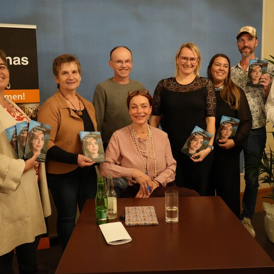 Eine Gruppe von Menschen steht hinter einem Tisch, lächelt und hält Bücher. Eine Frau in Pink hält einen Stift. Ein Mann in einem Hut hält ein Buch und ein anderer Mann ein Glas. Eine Topfpflanze steht rechts.