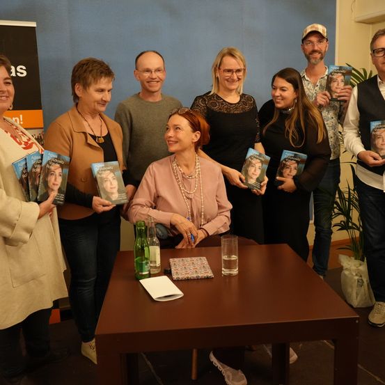 Eine Gruppe von Menschen, darunter Frauen mit Büchern, steht um einen Tisch herum. Sie scheinen bei einer Buchsignierstunde zu sein.