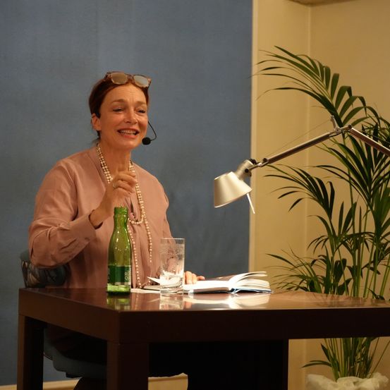 Eine Frau mit einer Brille auf dem Kopf sitzt an einem Tisch mit einem Buch und einem Glas Wasser. Sie trägt eine Halskette und einen Ring. Eine Lampe und eine Topfpflanze befinden sich an der Seite.