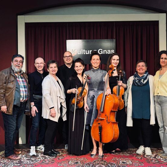 Eine Gruppe von Musikern posiert für ein Foto vor einem Hintergrund mit dem Wort Kultur Gnas. Sie halten Violinen und Celli und tragen lässige Kleidung.