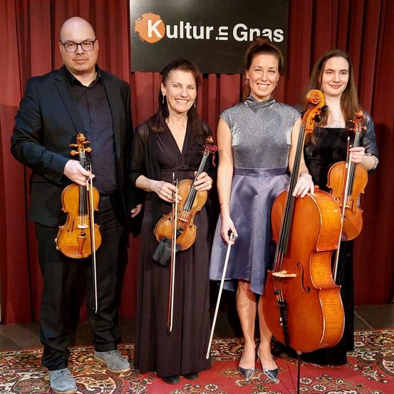 Vier Musiker stehen zusammen auf der Bühne, jeder hält eine Violine und Cello und lächelt vor einem roten Vorhang mit einem Kulture Gras-Schild.