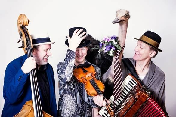 Three musicians and an ostrich pose for a photo, each holding a musical instrument. The man on the left plays the cello, the man in the middle plays the violin, and the man on the right plays the accordion. The ostrich has a bouquet of flowers on its neck.