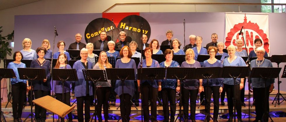 Ein Chor namens 'Comeditar Harmony' steht auf der Bühne, die Mitglieder tragen blaue Hemden und schwarze Hosen. Sie stehen hinter Notenständern, mit einem Hintergrund, der den Text 'Comeditar Harmony' zeigt.