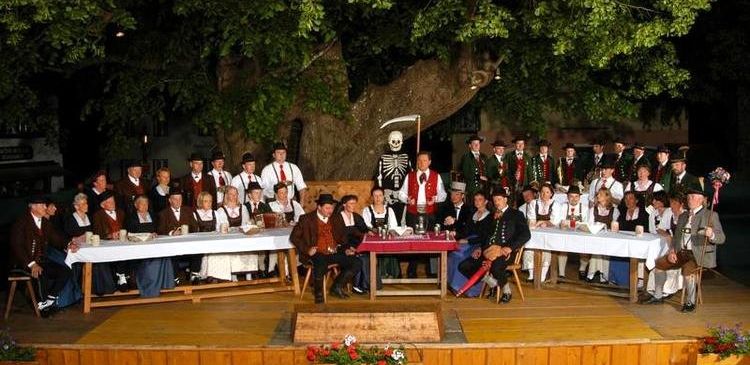 Eine Gruppe von Menschen in traditionellen Kostümen sitzt auf einem Bühnentisch, möglicherweise für eine Aufführung, mit einem Skelett, das eine Sense hält, dahinter.