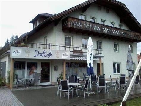 Die Vorderseite eines weißen Gebäudes mit Balkon hat ein Schild mit der Aufschrift Dorfcafe. Vor dem Gebäude stehen mehrere Stühle und Tische mit Schirmen.