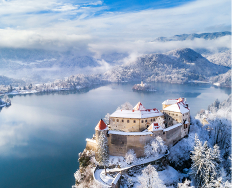Eine Luftaufnahme der Burg Bled in Slowenien, umgeben von einer verschneiten Landschaft und einem ruhigen See. Die Burg ist mit Schnee bedeckt, und Berge sind im Hintergrund.
