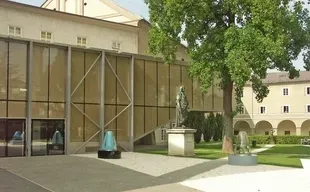 Ein modernes Gebäude mit Glaswänden und einem großen Baum davor. Eine Statue steht auf dem Rasen vor dem Gebäude. Eine Metallzaun und eine Glaswand befinden sich auf der linken Seite.