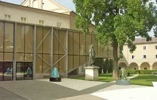Ein modernes Gebäude mit Glaswänden und einem großen Baum davor. Eine Statue steht auf dem Rasen vor dem Gebäude. Eine Metallzaun und eine Glaswand befinden sich auf der linken Seite.