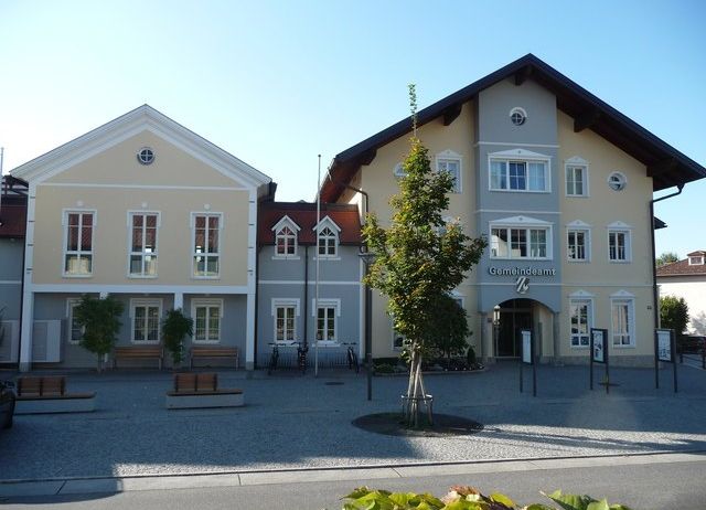 Das Bild zeigt ein Rathausgebäude mit vielen Fenstern, Bänken und geparkten Fahrrädern davor. In der Mitte des Gebäudes steht ein Baum.