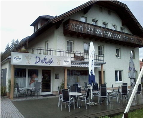 Ein weißes Gebäude mit Balkon und einem Schild, das Dorfcafe liest. Draußen gibt es Tische und Stühle mit Sonnenschirmen.