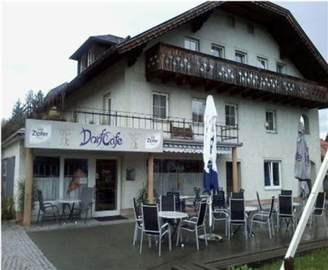 Ein weißes Gebäude mit dem Namen DorfCafe an der Vorderseite. Es hat einen Balkon, eine Terrasse mit Tischen und Stühlen und eine offene Tür.