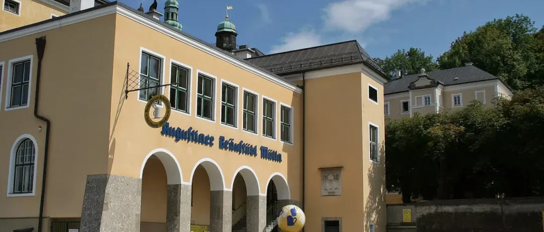 Ein gelbes Gebäude mit gewölbten Eingängen und blauem Schild, das 'Augstiner Reichenhall Museum' besagt. Ein großer farbenfroher Globus steht in der Mitte des Innenhofs.