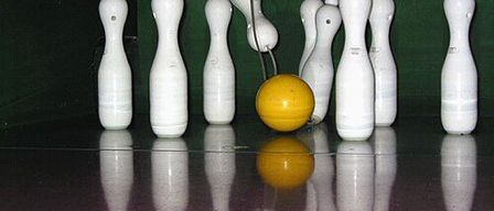 Bowlingkegel stehen auf einem glänzenden, dunklen Boden, mit einem gelben Bowlingball in der Mitte. Reflexionen erzeugen die Illusion mehrerer Kegel.