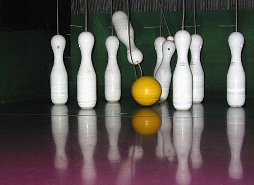 Bowlingkegel stehen auf einem glänzenden, dunklen Boden, mit einem gelben Bowlingball in der Mitte. Reflexionen erzeugen die Illusion mehrerer Kegel.