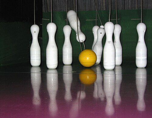 Eine Bowlingbahn mit einer gelben Kugel in der Mitte, umgeben von weißen Bowlingkegel, spiegelt sich auf einem glänzenden Boden wider.
