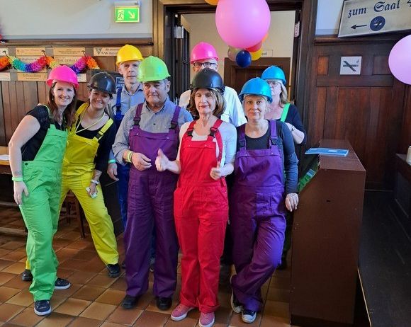 Eine Gruppe von Menschen in farbenfrohen Overalls und Helmen steht in einem Raum mit Luftballons, Fliesenboden und einem Holzschreibtisch.