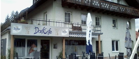 Ein weißes Gebäude mit Balkon, namens Dorfcafe, hat eine Terrasse mit Tischen, Stühlen und Sonnenschirmen. Bäume und Pflanzen sind in der Umgebung.
