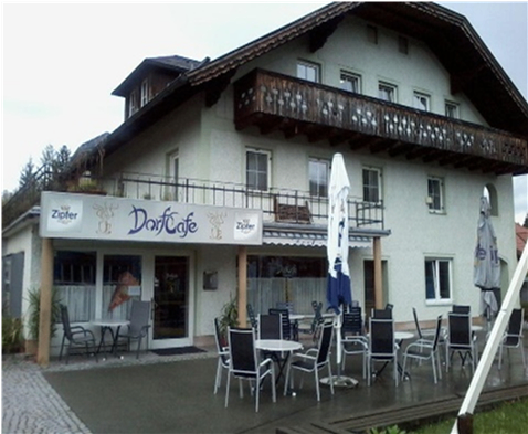 Ein weißes Gebäude mit Balkon, namens Dorfcafe, hat eine Terrasse mit Tischen, Stühlen und Sonnenschirmen. Bäume und Pflanzen sind in der Umgebung.