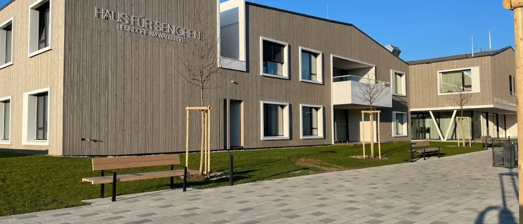 Ein modernes Gebäude mit der Aufschrift Haus für Senioren an der Wand, zwei Bänke links und ein Rasenbereich mit Pflanzen und Bäumen.