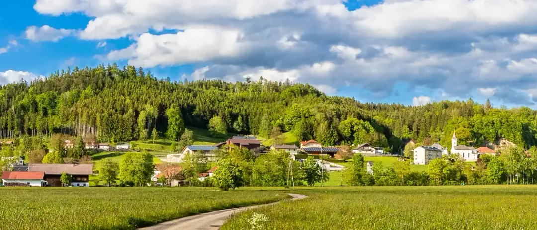 Eine ländliche Landschaft mit einem Dorf, das zwischen grünen Feldern liegt, umgeben von Bäumen und Bergen, unter einem teilweise bewölkten Himmel.