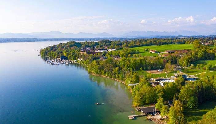Luftaufnahme eines Sees mit einer kleinen Stadt am Ufer und üppigen grünen Bäumen und Bergen in der Ferne.