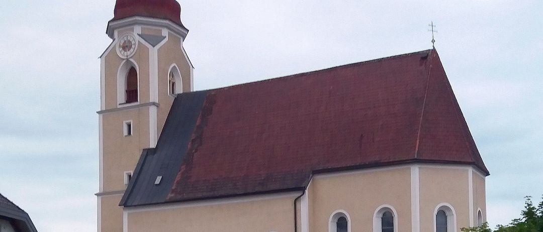 Eine beigefarbene Kirche mit rotem Dach und Turm, mit Rundbogenfenstern und einer Uhr, steht imposant in einer ländlichen Umgebung.