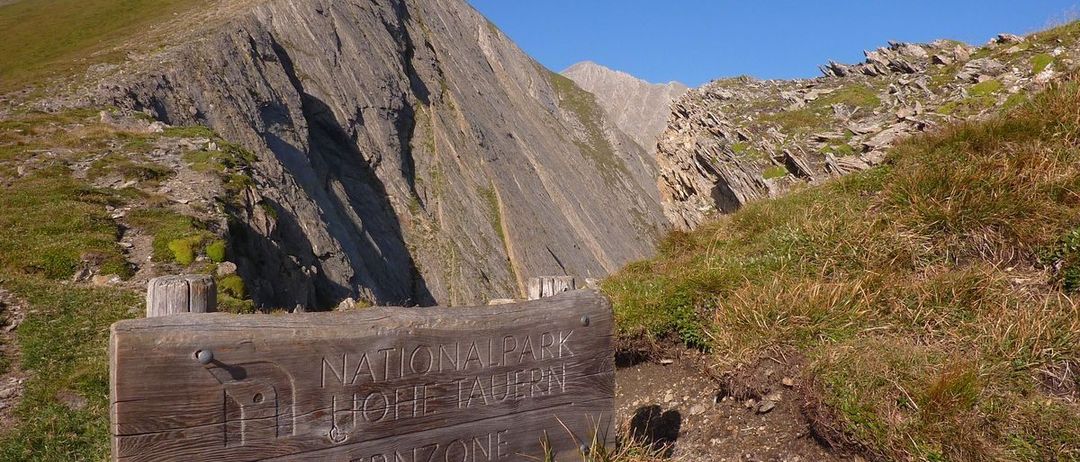 Ein hölzernes Wegweiser mit den Worten Nationalpark Hohe Tauern Kernzone steht auf einem Bergpfad. Hinter ihm erheben sich felsige Klippen, und das Gelände ist rau mit spärlicher Vegetation.