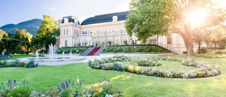 Ein großes Gebäude mit einem roten Teppicheingang, umgeben von üppigem Grün und einem Brunnen. Fahnen sind an der Fassade angebracht.