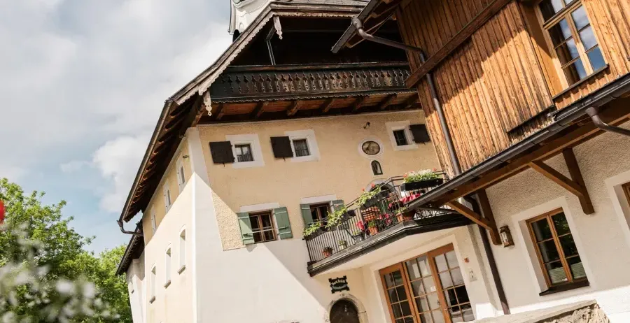 Ein traditionelles Gebäude mit einem Turm und einem Balkon mit Blumen, umgeben von Bäumen und gepflastertem Gehweg.