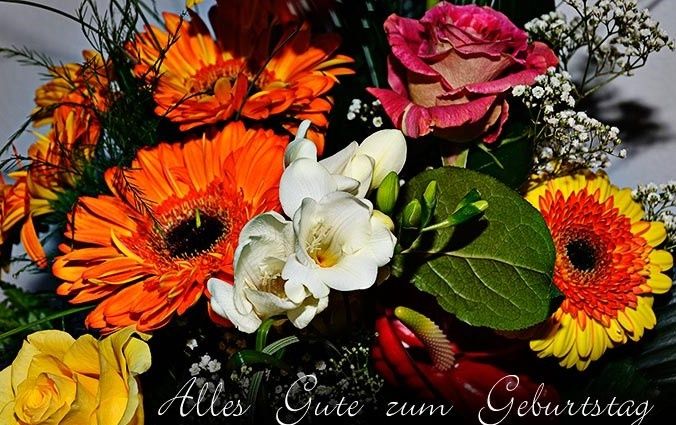 Ein Strauß bunter Blumen mit orangen und gelben Gänseblümchen, rosa und weißen Rosen und Babys Atmen. Der Strauß ist mit einer Nachricht in Deutsch, 'Alles Gute zum Geburtstag,' arrangiert.