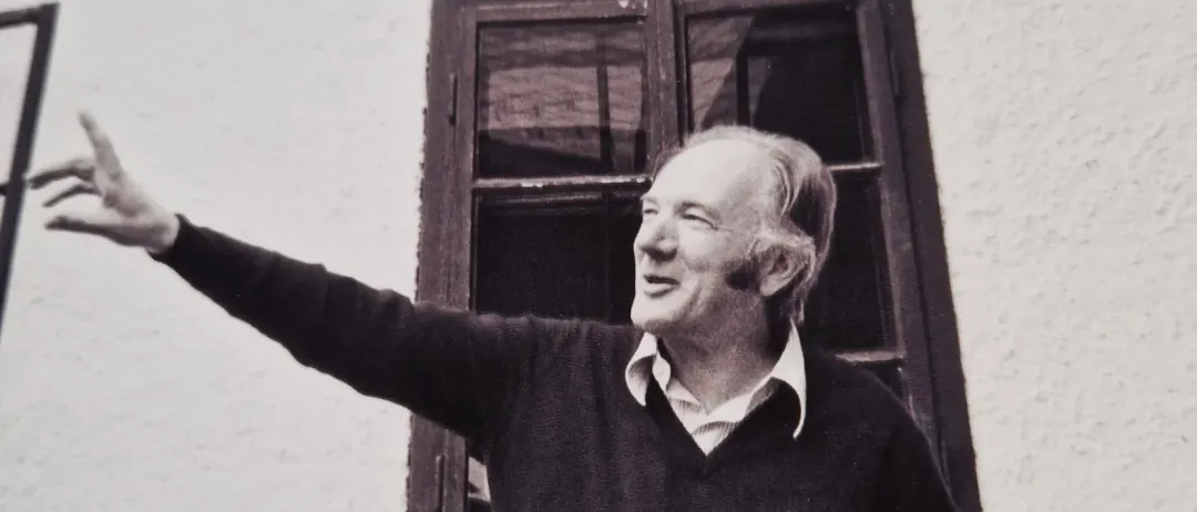 An elderly man with a white beard and a dark sweater stands outside a house and points to the side.